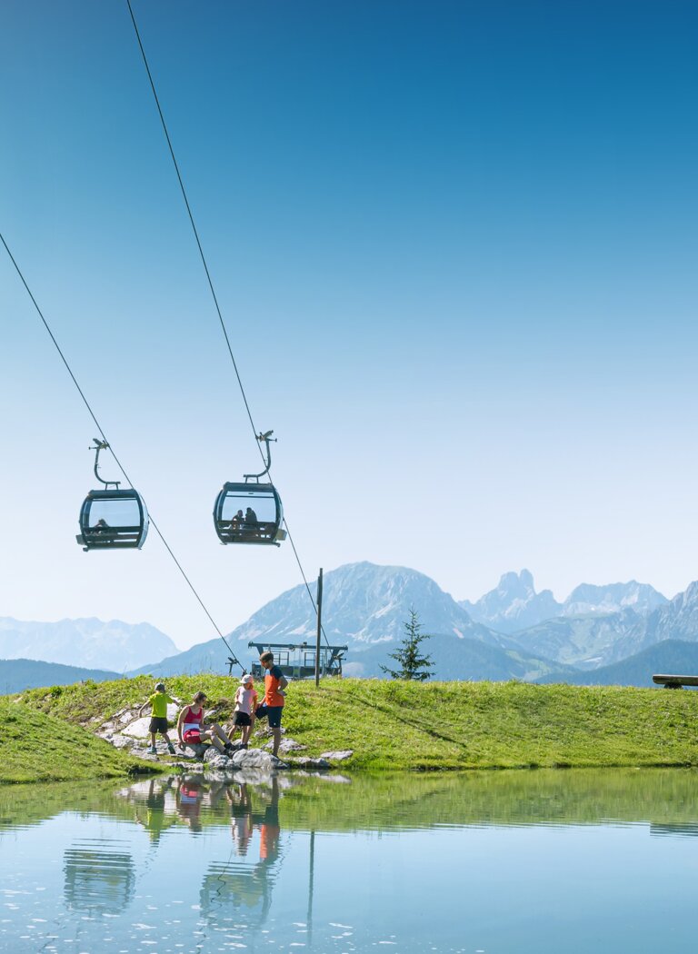 Familie am Ufer eines kleinen Bergsees unter einer Kabinenbahn, mit grünen Wiesen und Alpen im Hintergrund | © Ski amadé GmbH