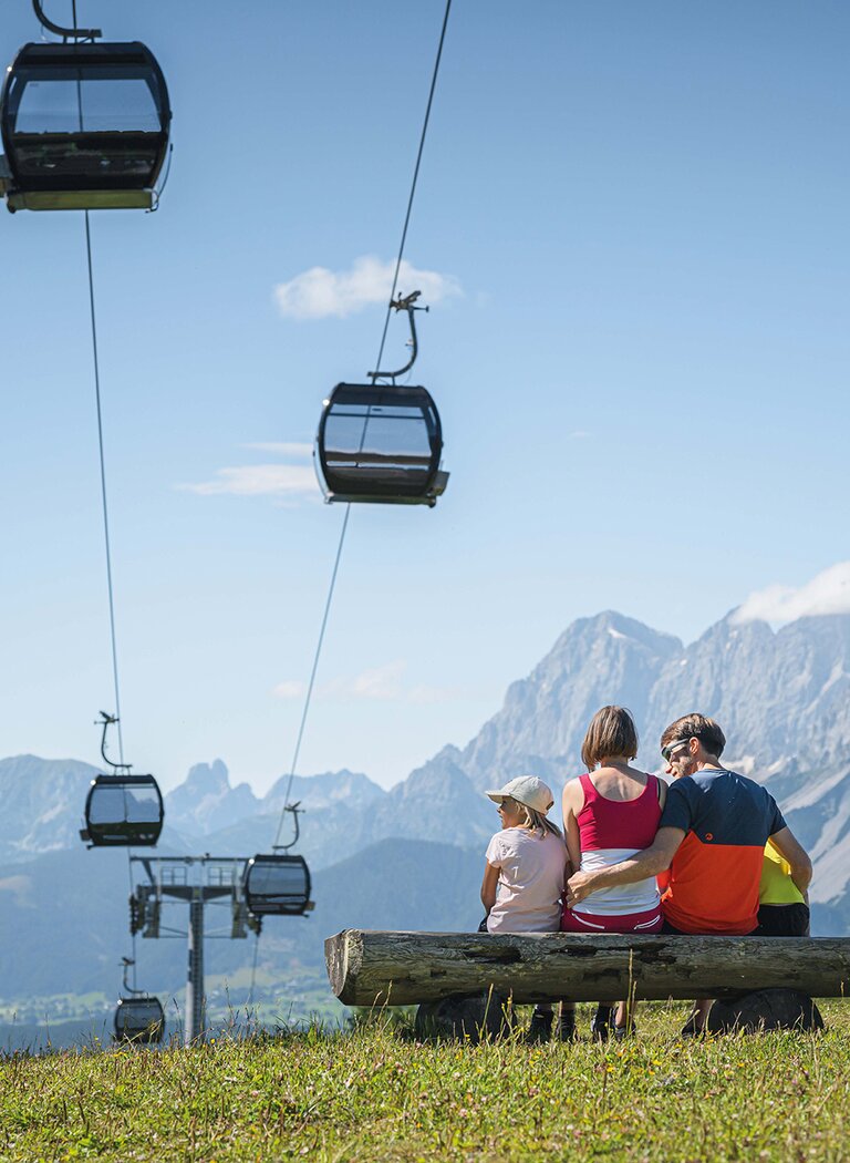 Family sitting on a bench overlooking gondolas and the impressive Dachstein mountain range. | © Ski amadé GmbH