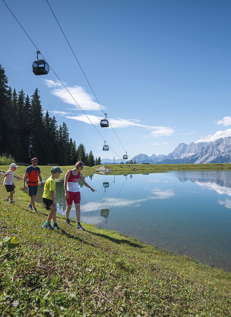 Family walking along a clear mountain lake, gondolas above, reflecting alpine peaks in calm water. | © Ski amadé GmbH