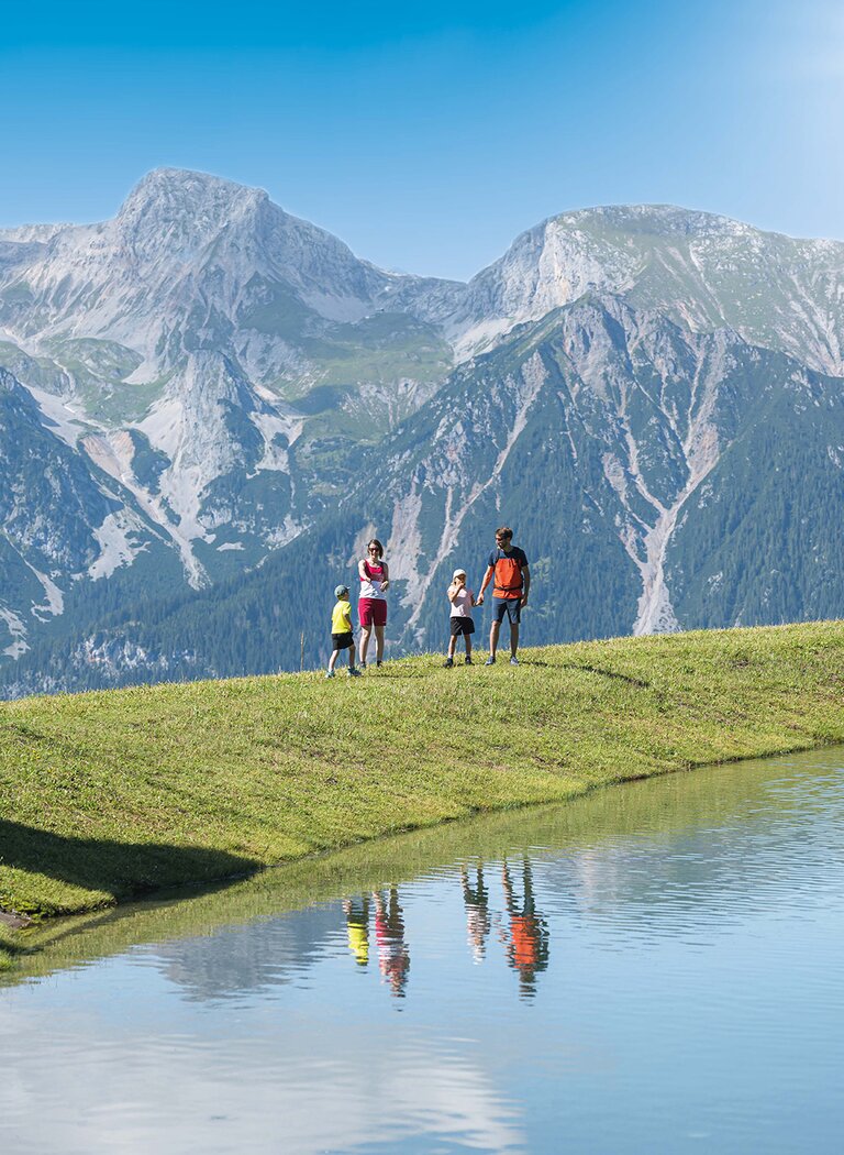 Familie wandert am klaren Bergsee, im Hintergrund imposantes Bergpanorama mit Spiegelung im Wasser. | © Ski amadé GmbH