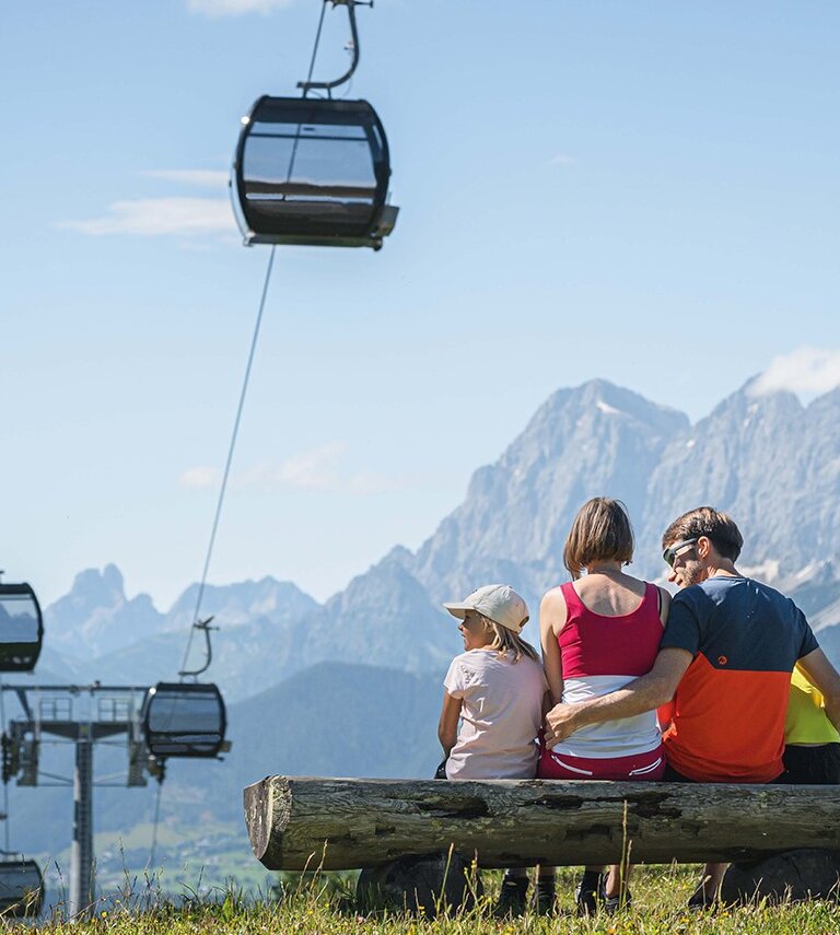 Familie sitzt auf Bank mit Blick auf Berge und Gondelbahn an einem sonnigen Sommertag. | © Ski amadé GmbH