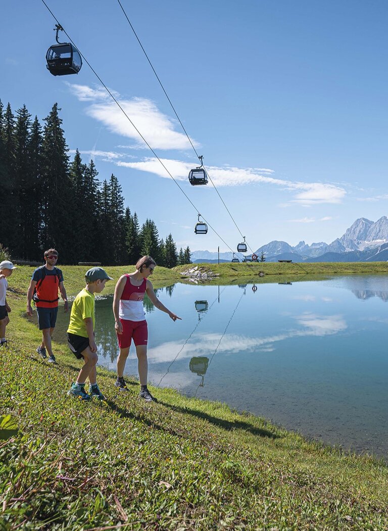 Familie wandert am klaren Bergsee entlang, mit Gondeln darüber und Bergpanorama im Hintergrund unter blauem Himmel. | © Ski amadé GmbH