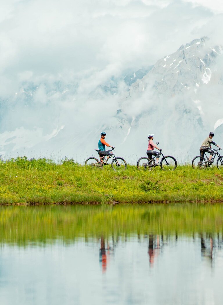 Drei Personen auf E-Bikes fahren am See entlang, im Hintergrund steile, wolkenverhangene Berge und Wasserspiegelung. | © Ski amadé GmbH