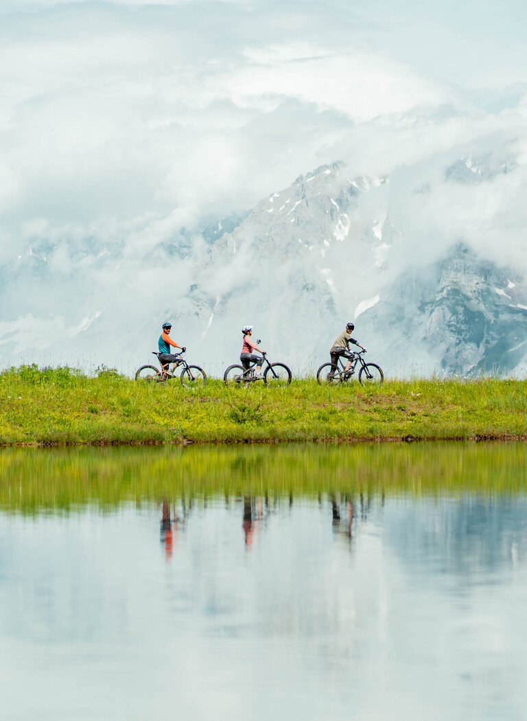 Three cyclists ride along a mountain lake, misty peaks in background and soft reflection on water surface. | © Ski amadé GmbH