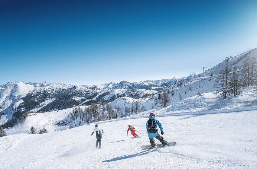 Drei Wintersportler – zwei Skifahrer und ein Snowboarder – fahren bei Sonne über breite Pisten mit Bergblick in Ski amadé. | © Ski amadé GmbH