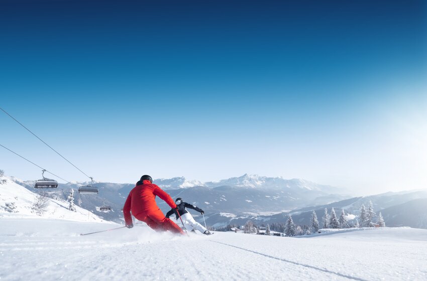 Zwei Skifahrer in Rot und Schwarz fahren schwungvoll auf einer leeren Piste mit Blick auf das verschneite Tal in Ski amadé. | © Ski amadé GmbH