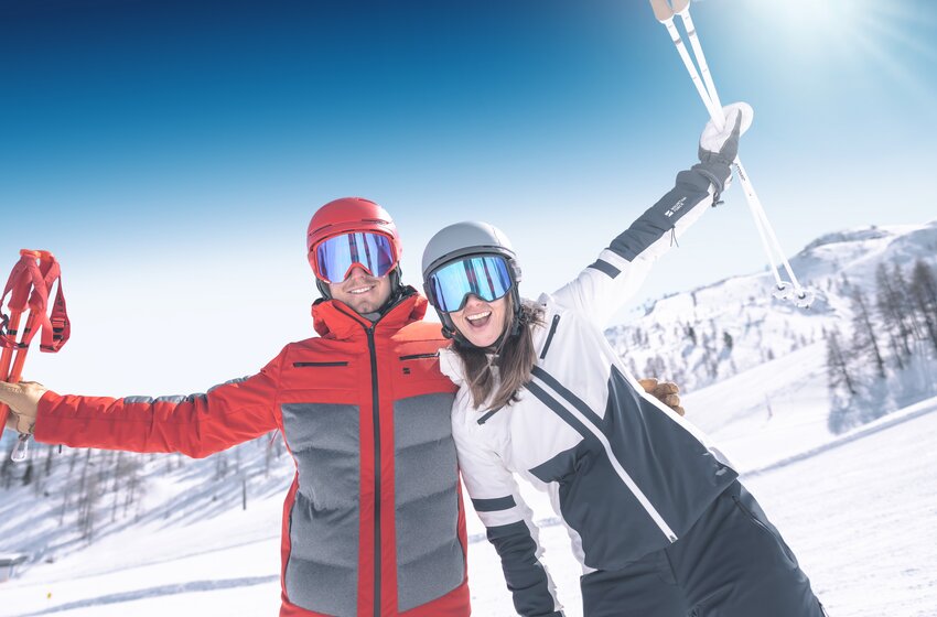 Zwei Skifahrer stehen lachend nebeneinander im Schnee, Arme hochgestreckt, mit Helm und Brille vor sonniger Winterkulisse. | © Ski amadé GmbH