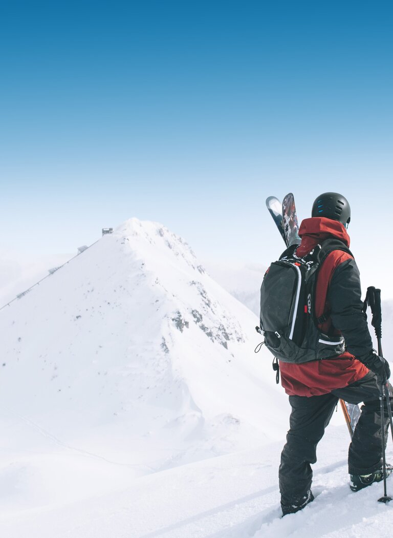 Two winter sports enthusiasts with gear and helmets stand on a summit ridge, facing a snowy slope in Ski amadé backcountry. | © Ski amadé GmbH