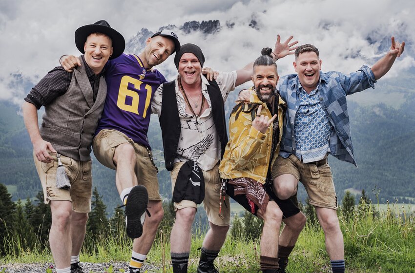 Five men in lederhosen pose arm in arm on a grassy hillside with rocky mountains and low clouds behind them. | © Angelo Lair