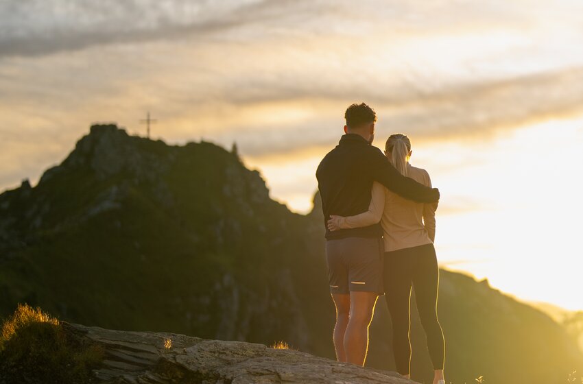 Paar steht eng umschlungen auf Felsen am Berggipfel bei Sonnenuntergang, Berge und Himmel im Hintergrund | © Gasteiner Bergbahnen AG