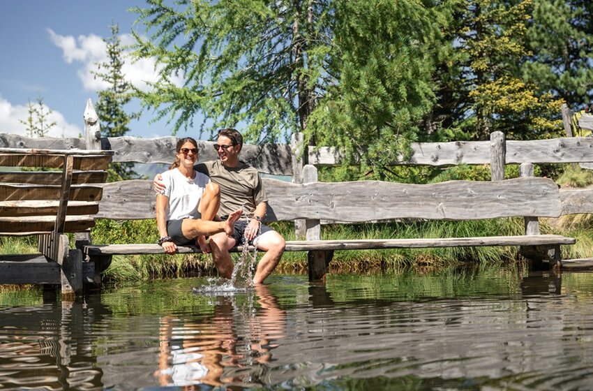 Paar sitzt auf Holzbank am Teich und hält Füße ins Wasser, Spritzer sichtbar, umgeben von Bäumen und Zaun | © Gasteiner Bergbahnen AG