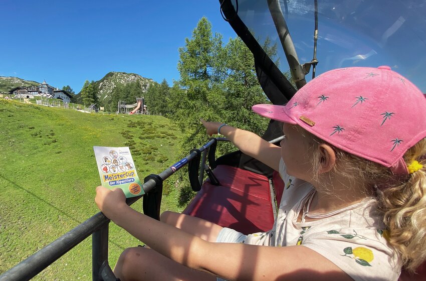 Mädchen mit pinker Kappe hält MeisterCup-Karte und zeigt beim Bergabfahren mit der Gamskogelbahn auf den Abenteuerspielplatz. | © Zauchensee Liftgesellschaft Benedikt Scheffer GmbH
