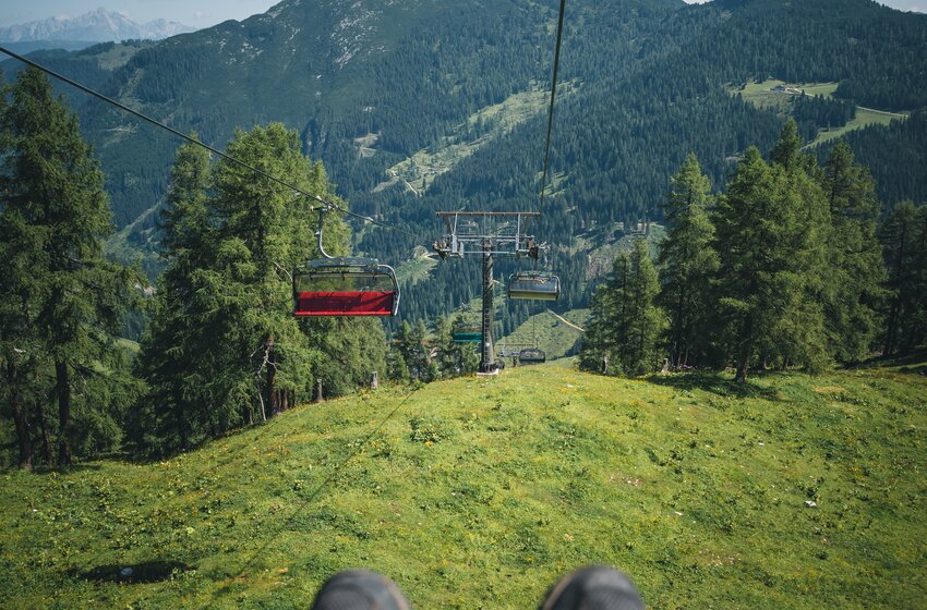 Talfahrt mit dem Sessellift, Wanderschuhe im Vordergrund, darunter grüne Almwiese und dahinter bewaldete Berglandschaft. | © Zauchensee Liftgesellschaft Benedikt Scheffer GmbH