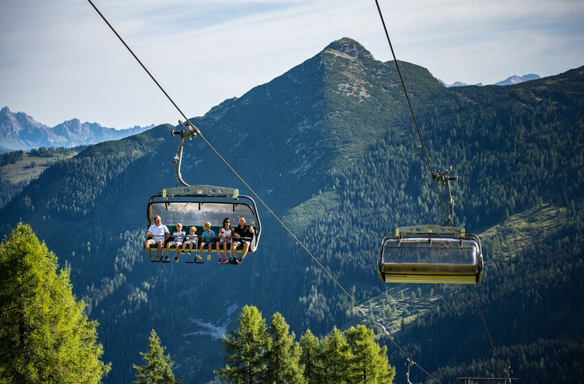 A family rides a chairlift in front of forested mountains on a sunny day above the alpine slope. | © Zauchensee Liftgesellschaft Benedikt Scheffer GmbH