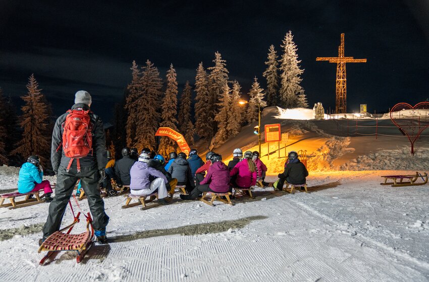 Menschen auf Schlitten sitzen bei Nacht vor beleuchteter Rodelbahn mit Kreuz und verschneiten Bäumen | © Zauchensee Liftgesellschaft Benedikt Scheffer GmbH
