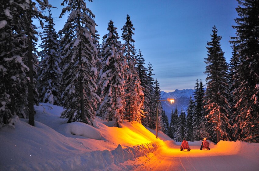 Zwei Rodler fahren bei Dämmerung auf beleuchteter Schneepiste durch verschneiten Wald mit Bergblick | © Zauchensee Liftgesellschaft Benedikt Scheffer GmbH